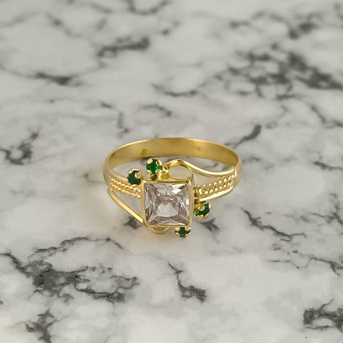 Anillo Emperatriz en oro laminado 18k con circón cuadrado y piedras verdes sobre fondo de mármol.