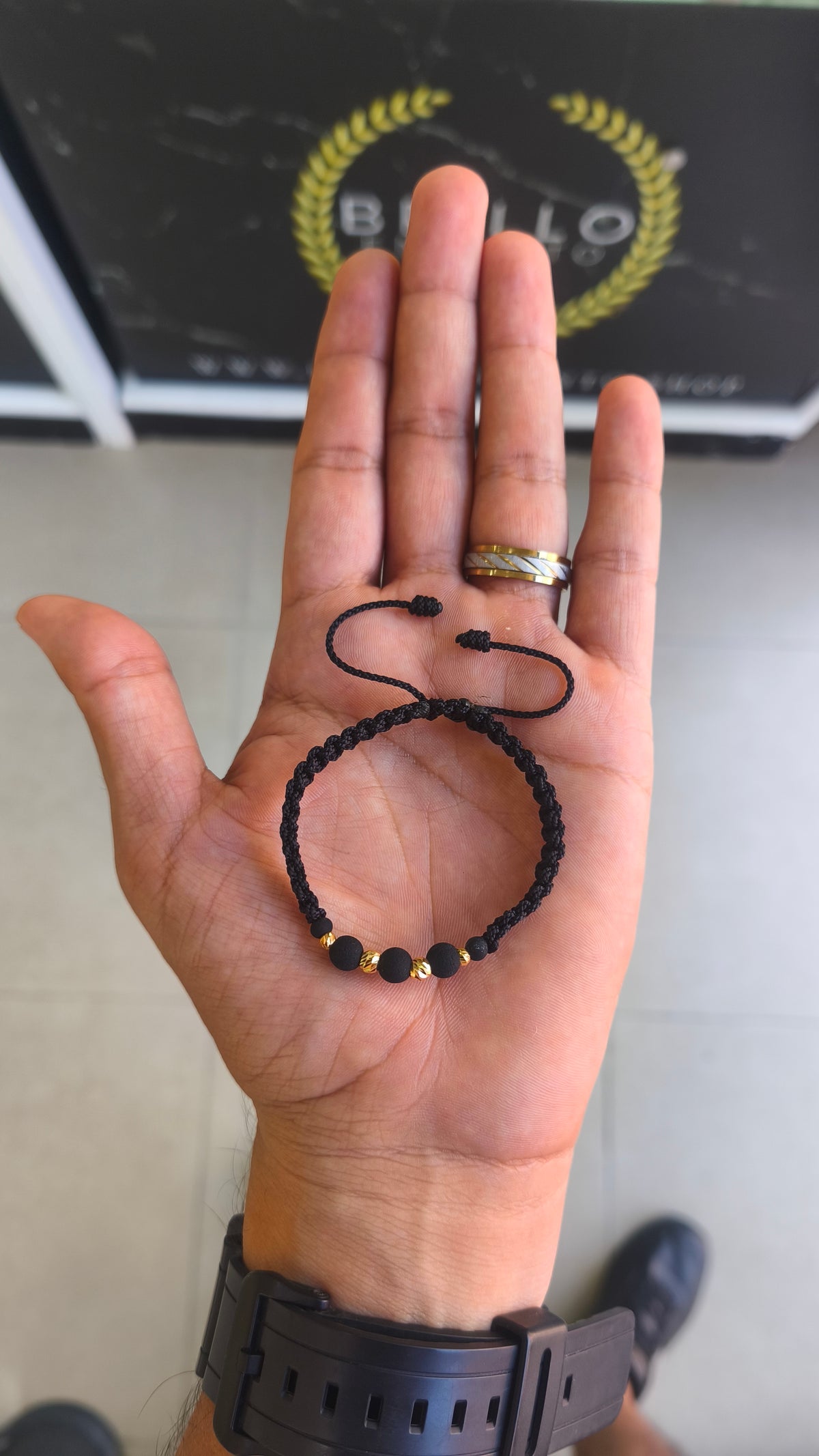 Pulsera unisex de hilo negro con cuentas negras y balines de oro 18K, sostenida frente al logo de Brillo Encanto.