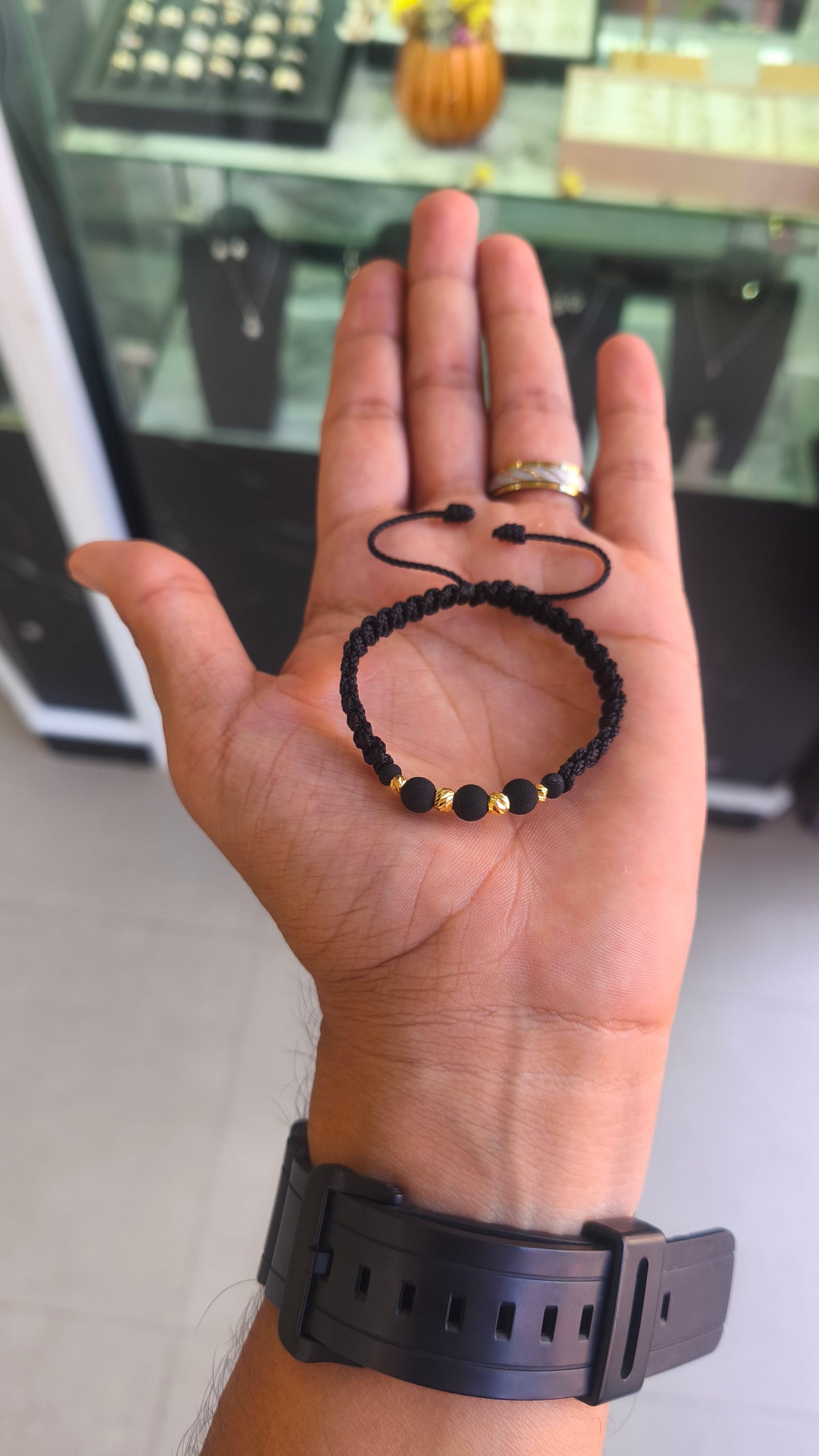 sobre la palma de la mano: Pulsera de balines de oro 18K y cuentas negras sobre la palma de una mano, con tejido negro y cordones ajustables.