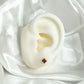 Arete pequeño de bebé con diseño de trébol rojo en oro laminado 18K, colocado en oreja de maniquí blanco, ideal para niñas recién nacidas.