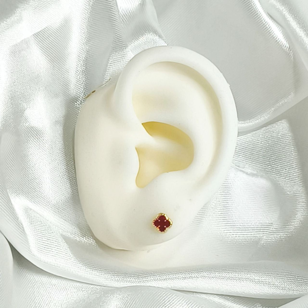 Arete pequeño de bebé con diseño de trébol rojo en oro laminado 18K, colocado en oreja de maniquí blanco, ideal para niñas recién nacidas.